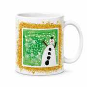 A white ceramic mug featuring a festive winter design. The mug is decorated with a glittery gold border surrounding a square illustration of a snowman on a green snowy background.